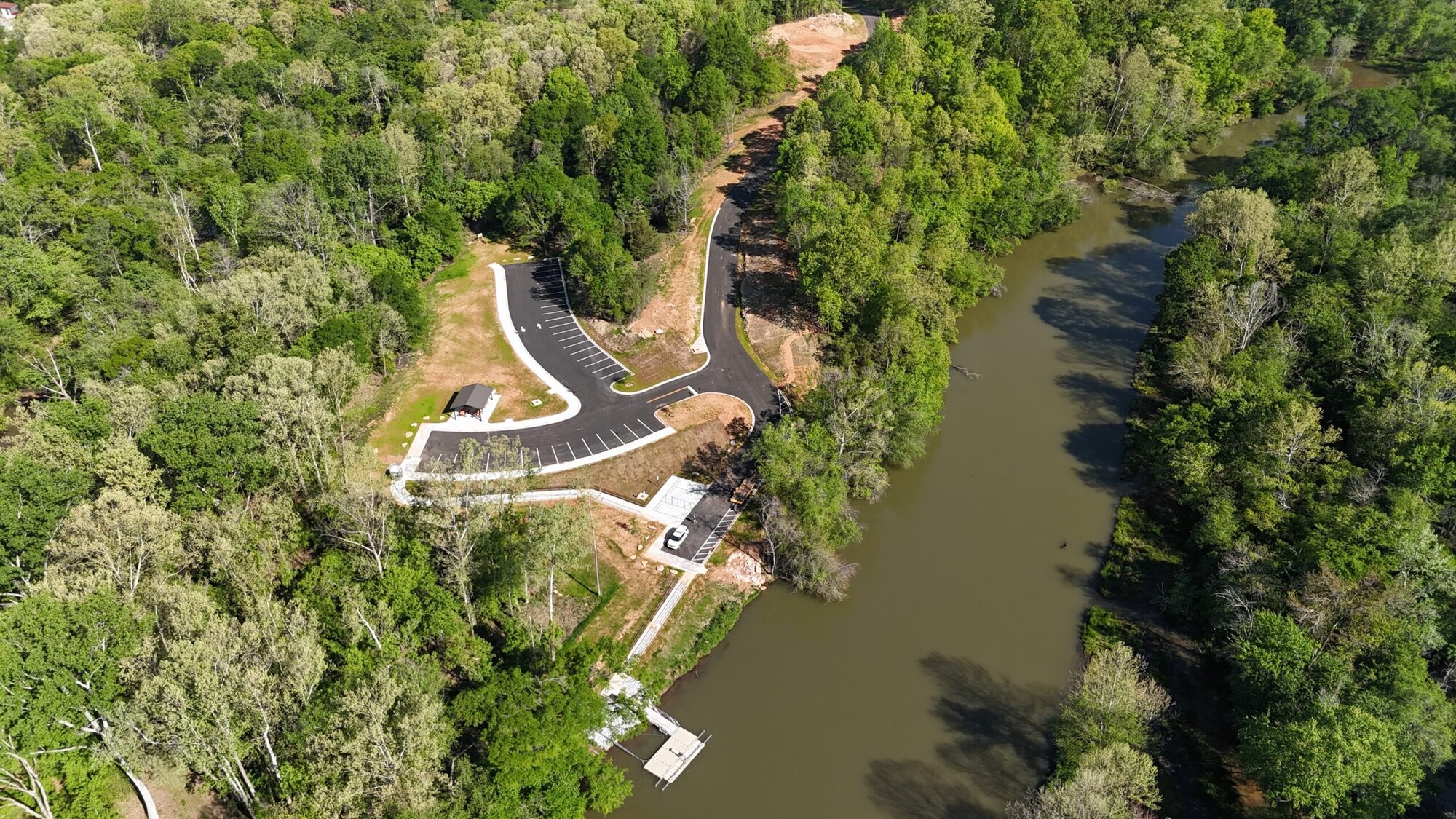 This is an aerial (drone-style) view of a riverside park or access point surrounded by dense forest. Key elements visible: * **River**: On the right side, a calm, slightly murky river runs vertically through the frame, bordered by thick trees on both banks. * **Boat access area**: Near the bottom center-right, there’s a small dock or launch platform extending into the water, likely for kayaks or small boats. * **Parking lot**: On the left side of the river, a newly paved parking area with marked spaces curves along the landscape. It appears lightly used or empty. * **Access road**: A narrow road winds down from the top center toward the parking area, cutting through the forest. * **Shelter structure**: A small pavilion or covered structure sits near the parking lot, likely for picnics or resting. * **Cleared land**: Some areas around the parking lot and road look recently cleared or developed, with exposed soil contrasting the surrounding greenery. * **Dense forest**: The majority of the scene is covered in lush, green trees, suggesting a natural or protected area. Overall, it looks like a newly developed recreational access point—possibly for boating, fishing, or nature visits—integrated into a wooded riverside environment.